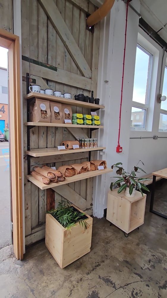 Interior corner with wooden retail shelves holding coffee bags, jars and pastries beside a planter